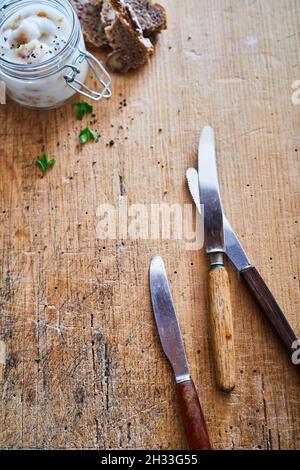 Vertikale Draufsicht Satz von rostfreien Messern auf einem Tisch mit einem Glasglas mit Schmalz-Aufstrich und Brot Stockfoto