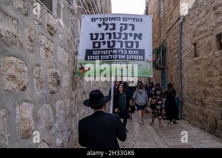 Ein religiöser Jude hält ein Schild mit der Aufschrift „Ein glückseliges Leben ist nur ohne Internet und Filme“, während er durch eine Gasse im jüdischen Viertel in der Altstadt von Jerusalem, Israel, geht. Haredi-Gemeinden meiden routinemäßig moderne Technologien wie Internet und Smartphones. Ihre Argumentation ist, dass die immense Menge an Informationen korrumpieren kann, und mit der Fähigkeit, das Internet ohne Beobachtung durch die Gemeinschaft zu nutzen, kann zu Individuation führen. Stockfoto