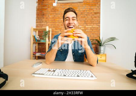 Vorderansicht eines lachenden hungrigen jungen Mannes, der Hamburger mit Rindfleisch aus dem Fast-Food-Restaurant hält, der mit einer kabellosen Tastatur an der Rezeption sitzt Stockfoto