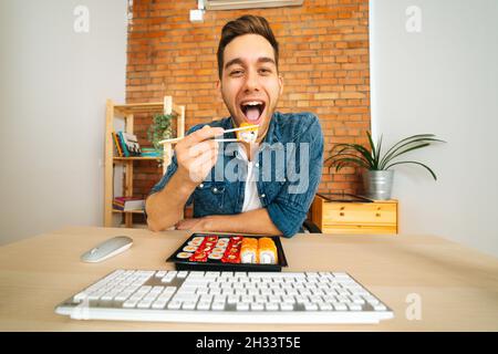 Vorderansicht eines fröhlichen, hübschen jungen Mannes, der Sushi isst und Informationen vom Monitor liest und die Kamera anschaut. Stockfoto