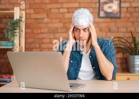 Porträt von Angst Angst Angst männlichen Verschwörungstheoretiker tragen Aluminiumfolie schützen Gehirn beobachten schockierte Online-Video-Inhalte mit Laptop-Computer. Stockfoto