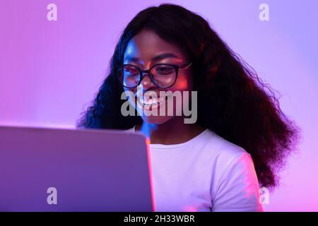 Glückliche junge schwarze Frau in einer Brille mit Laptop-pc für Online-Arbeit oder Studien in Neonlicht. Fröhliche afroamerikanische Dame mit Remote-Video-Conferen Stockfoto