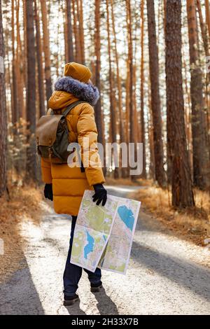 Junge Wanderin in einer warmen orangefarbenen Jacke. Eine Frau spaziert in einem herbstlichen Kiefernpark mit einem Rucksack. Wandern im Wald in der Herbstsaison. Rückansicht. V Stockfoto