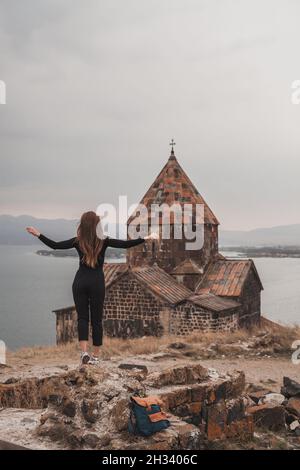 Eine junge Frau mit langen braunen Haaren in schwarzer Kleidung steht in der Nähe der alten Steinkirche am Meer. Reisende spazieren in der Nähe der alten Kapelle in Armenien. Stockfoto