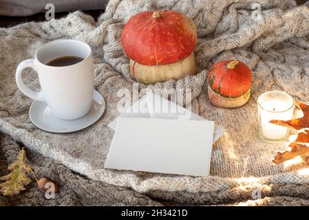Gemütliche Herbstfrühstückszene. Leere Grußkarte Mockup und Umschlag mit einer Tasse Kaffee oder Tee, Kerze, Kürbisse.Plaid Wolle Hintergrund.Fall, danke Stockfoto
