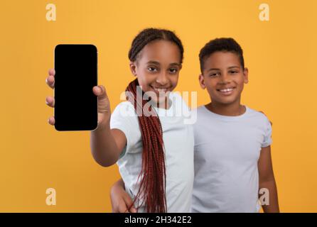 Niedliche afroamerikanische Kinder Teenager Junge und Mädchen zeigt brandneues Smartphone mit leerem Bildschirm, umarmt und lächelt an der Kamera, zeigt schöne Unterhaltung Stockfoto