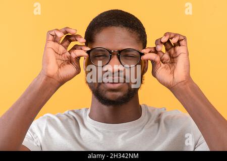 afroamerikanischer Mann mit schlechter Sehkraft, der eine Brille trägt und Augen schielt, während er durch eine Brille auf die Kamera blickt, gelber Studiohintergrund. Gesund Stockfoto