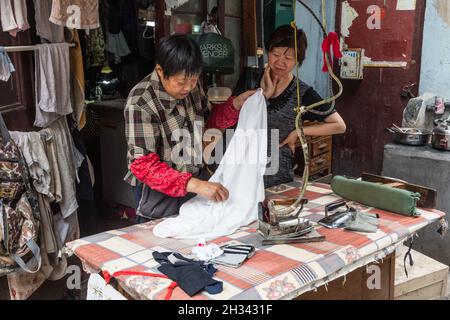 Chinesische Frauen bügeln Kleidung in einem offenen Schneiderladen am Straßenrand im alten Teil von Shanghai, China. Stockfoto