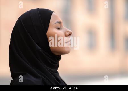 Außenprofil Porträt Einer Ruhigen Jungen Muslimischen Frau In Schwarz Hijab Stehend Mit Geschlossenen Augen, Seitenansicht Aufnahme Von Friedlicher Religiöser Islamischer Kleidung Stockfoto