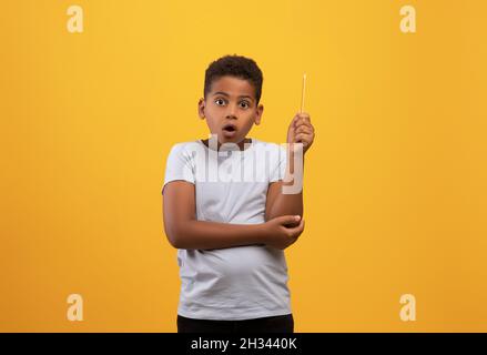 Staunender schwarzer Schuljunge, der Bleistift auf gelben Studiohintergrund hob, schlaues Kind mit schockierter Gesichtsausdruck, das eine kreative Lösung hat und die eu zeigt Stockfoto