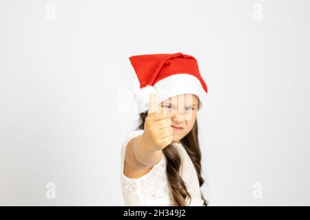 Porträt eines Mädchens in weißem Kleid und trägt roten Weihnachtsmann Hut gibt Daumen nach oben, isoliert auf weißem Hintergrund mit Kopierraum Stockfoto