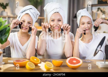 Fürsorgliche Mutter mit zwei niedlichen Töchtern, die lächeln und die Kamera betrachten, während sie schwarze Kollagenflecken in den Händen hält. Kosmetik für Schönheitsbehandlungen zu Hause. Hautpflege und Mutterschaft Konzept. Stockfoto