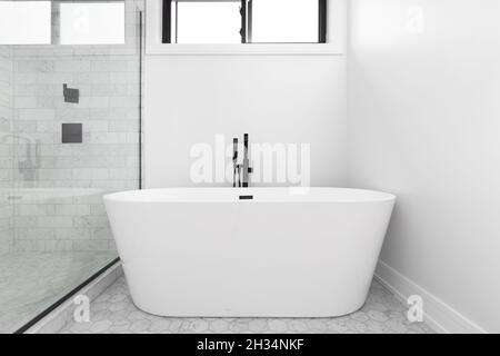 Eine renovierte, eigenständige Badewanne mit schwarzem Wasserhahn, kleinen sechseckigen Fliesen auf dem Boden und einer Dusche mit Glaswänden und großen Marmorfliesen. Stockfoto