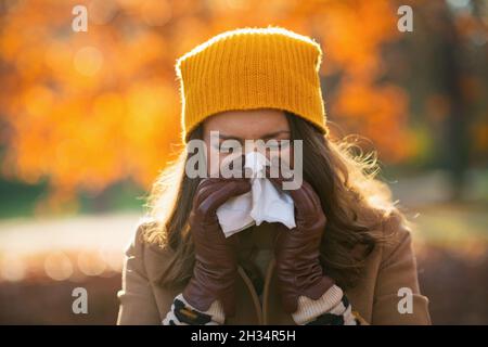 Hallo Herbst. Elegantes Weibchen in beigem Mantel und orangefarbenem Hut mit Serviette, die im Herbst im Stadtpark die Nase weht. Stockfoto