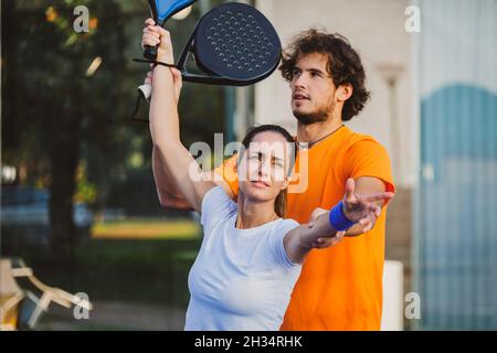 Der junge Lehrer überwacht den Padel-Unterricht für seine Schülerin - Coach lehrt Mädchen, wie man Padel auf dem Tennisplatz im Freien spielt Stockfoto