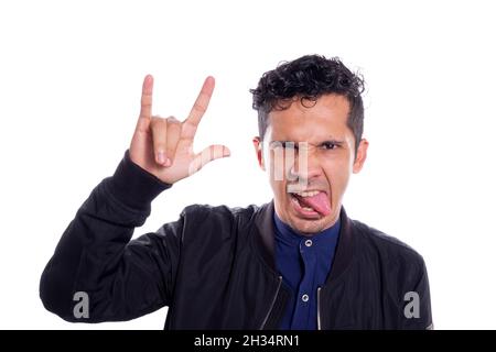 Mann macht Rocker Geste, auf weißem Hintergrund. Junger erwachsener junger Mann, der seine Zunge mit Rocker-Pose, Hand mit Rock and Roll-Zeichen hervorsticht. Stockfoto