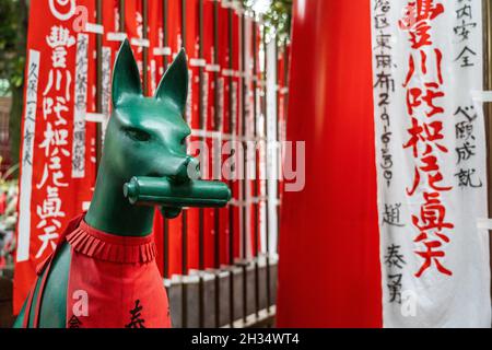 Eine Statue der Inari Kitsune oder Fuchsgöttin, die ein rotes Lätzchen trägt und von Nobori-Bannern umgeben ist, am Toyokawa Inari Betsuin Tempel in Asakusa, Tokio, Japan. Der buddhistische Tempel ist Teil der Soto Zen Sekte und beherbergt die Gottheit Toyokawa Dakinishinten, aber auch bekannt für die Tausenden von Fuchsstatuen. Stockfoto