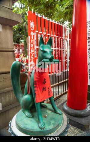 Eine Statue der Inari Kitsune oder Fuchsgöttin, die ein rotes Lätzchen trägt und von Nobori-Bannern umgeben ist, am Toyokawa Inari Betsuin Tempel in Asakusa, Tokio, Japan. Der buddhistische Tempel ist Teil der Soto Zen Sekte und beherbergt die Gottheit Toyokawa Dakinishinten, aber auch bekannt für die Tausenden von Fuchsstatuen. Stockfoto