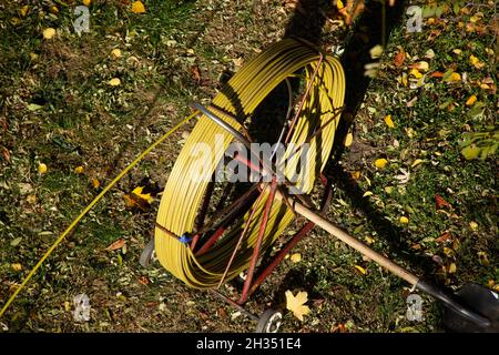 Ein großer Strang gelber Draht, gewickelt auf einer eisernen Trommel vor einem Hintergrund aus grünem Gras und gelben Herbstblättern. Ausrüstung für die Reinigung von Rohrleitungen, Stockfoto