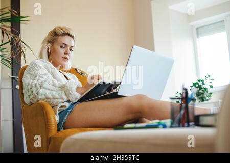 Ansicht von unten einer kaukasischen Frau, die Online-Lektionen hört. Bequem auf der Couch stehend, mit ausgestreckten Beinen auf dem Tisch, aufmerkend Stockfoto