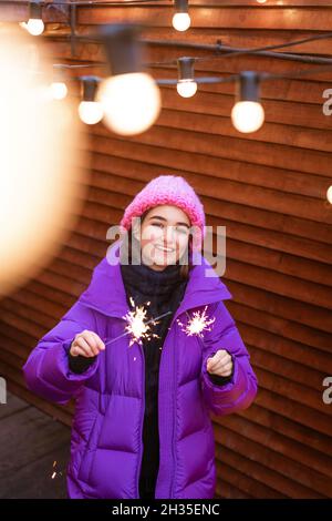 Kaukasisches Mädchen hat Spaß im Winter auf der Straße mit Wunderkerzen in der Hand lächelt vor Glück, während sie in einer warmen Fliederjacke und Strickmütze steht. Helle Funken aus den Lichtern Stockfoto