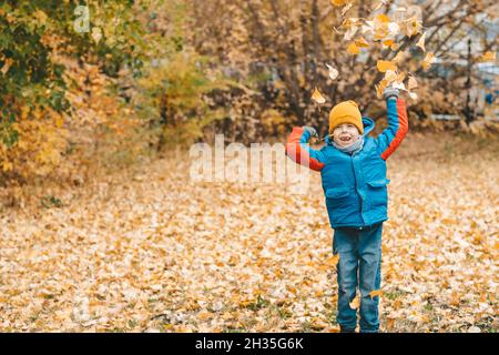 Junge in einer blauen Jacke, streut Blätter in einem Herbstpark. Das Kind freut sich über die Herbstblätter. Frohe Kindheit. Leuchtend gelbe Jacke und Blätter. Wa Stockfoto