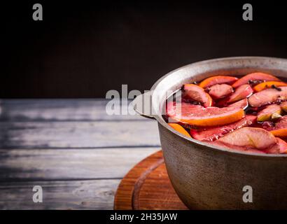 Großer Wasserkocher Topf mit traditionellen heiß dampfenden Weihnachtsgetränk Glühwein rot gewürzt kocht mit Äpfeln und Zitrusfrüchten auf schwarzem Hintergrund. Weihnachtsfeier. Stockfoto