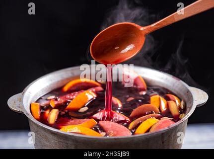 Großer Wasserkocher Topf mit traditionellen heiß dampfenden Weihnachtsgetränk Glühwein rot gewürzt kocht mit Äpfeln und Zitrusfrüchten mit Holzlöffel auf schwarzem Hintergrund Stockfoto