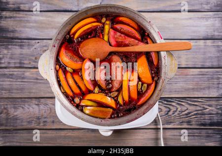 Großer Wasserkocher Topf mit traditionellen heiß dampfenden Weihnachtsgetränk Glühwein rot gewürzt kocht mit Äpfeln und Zitrusfrüchten mit Holzlöffel auf schwarzem Hintergrund Stockfoto