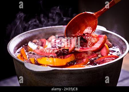 Großer Wasserkocher Topf mit traditionellen heiß dampfenden Weihnachtsgetränk Gluhwein Glühwein rot gewürzten Wein kocht mit Äpfeln und Zitrusfrüchten mit Holzlöffel auf schwarz b Stockfoto