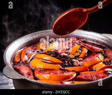 Großer Wasserkocher Topf mit traditionellen heiß dampfenden Weihnachtsgetränk Glühwein rot gewürzt kocht mit Äpfeln und Zitrusfrüchten mit Holzlöffel auf schwarzem Hintergrund Stockfoto