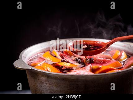 Großer Wasserkocher Topf mit traditionellen heiß dampfenden Weihnachtsgetränk Gluhwein Glühwein rot gewürzten Wein kocht mit Äpfeln und Zitrusfrüchten mit Holzlöffel auf schwarz b Stockfoto