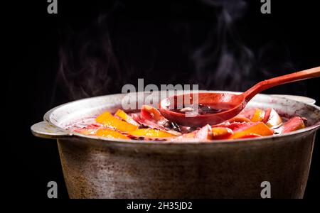 Großer Wasserkocher Topf mit traditionellen heiß dampfenden Weihnachtsgetränk Glühwein rot gewürzt kocht mit Äpfeln und Zitrusfrüchten mit Holzlöffel auf schwarzem Hintergrund Stockfoto