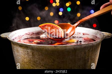 Großer Wasserkocher Topf mit traditionellen heiß dampfenden Weihnachtsgetränk Glühwein rot gewürzt kocht mit Äpfeln und Zitrusfrüchten mit Holzlöffel auf schwarzem Hintergrund Stockfoto