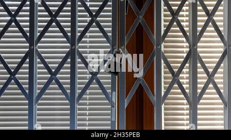 Klappgitter aus Stahl vor einem geschlossenen Fensterladen, der vom Raum aus sichtbar ist. Das Fenster von außen ist teilweise schattig und teilweise in der Morni Stockfoto