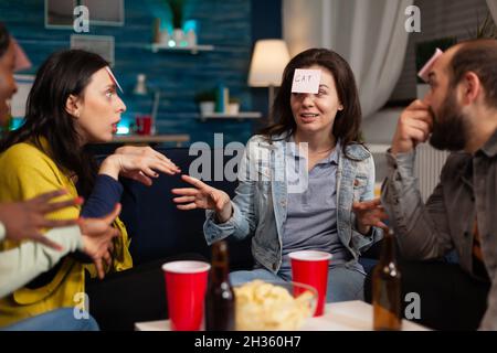 Porträt einer glücklichen Frau mit Stichnoten auf der Stirn spielen erraten, wer Spiel genießen Zeit mit Freunden Geselligkeit zusammen während wekeend Party zu Hause verbringen. Freundschaftskonzept Stockfoto