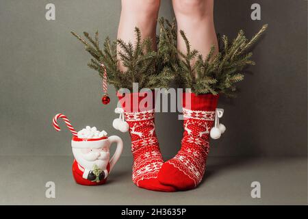 Weibliche Beine in roten Wollsocken mit Ornamenten, minimal verzierten Tannenzweigen und einer Tasse Kakao mit Sumpflaibchen, Zuckerrohr auf grünem Hintergrund. Cre Stockfoto