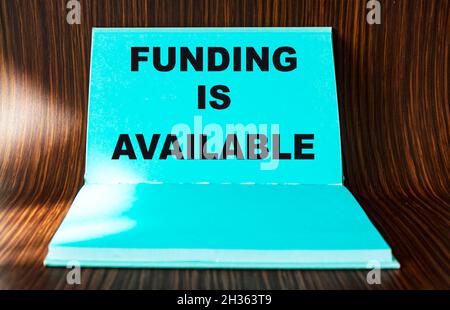 Es stehen Fördermittel zur Verfügung, der Text wird auf einen grünen Notizblock und einen Holztisch geschrieben. Geförderte Finanzierung Business Start-up-Projekte Konzept. Stockfoto