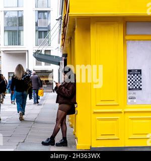 Junge ethnische Frau, die mit dem Mobiltelefon gegen das Gelbe Gebäude anstöhnen muss, während die Menschen London, England, passieren Stockfoto