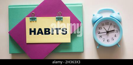 Das Wort Gewohnheit steht auf einem Aufkleber, der auf farbigen Tagebüchern liegt, neben einem Wecker auf weißem Hintergrund Stockfoto