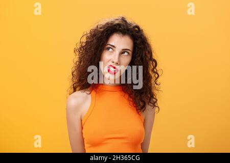 Stilvolle heiße dunkelhaarige Frau mit Locken und rotem Lippenstift in trendigen Cropped Top Blick auf die obere linke Ecke fasziniert und neugierig klemmende Zähne Stockfoto