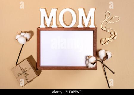 Festliches Layout für den Muttertag mit Rahmen, weißen Buchstaben MAMA, diy-Geschenkbox und Baumwollcobs auf beigem Hintergrund. Modisch monochrom. Modell. Blick von Stockfoto