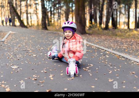 Kindermädchen in einem rosa Helm und vier Rädern Rollen sitzt auf dem Asphalt und lächelt. Herbst Outdoor-Aktivitäten Stockfoto