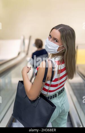 Junge, schöne Frau in weißer Maske und Handtasche, die über die Rolltreppe läuft und auf die Kamera schaut Stockfoto