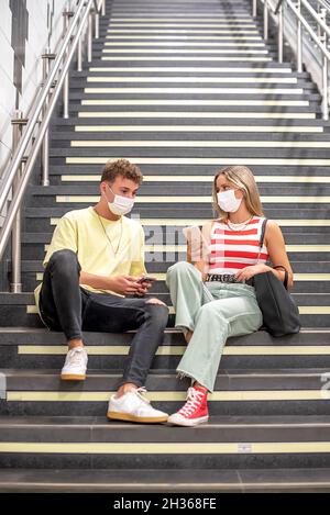 Ein junger Mann und eine Frau, die Masken tragen, sitzen auf der Treppe einer U-Bahnstation und unterhalten sich mit ihren Handys in der Hand Stockfoto