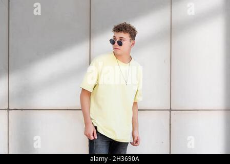 Junger Mann im gelben T-Shirt posiert mit seiner Sonnenbrille auf grauem Betongrund im Freien Stockfoto