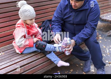 Ein Mann legt Knieschoner auf die Beine eines kleinen Mädchens, bevor er Rollschuhlaufen geht. Vatertag, Familie Stockfoto