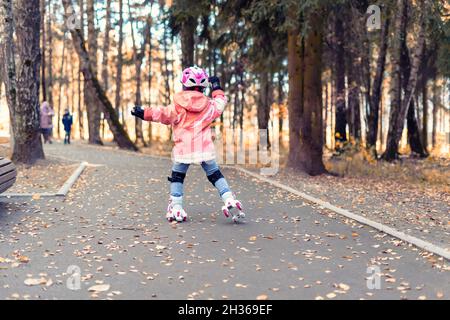 Kid girl in einem rosa Helm reitet in einem Herbstpark auf vier Rädern Rollen. Herbst Outdoor-Aktivitäten Stockfoto