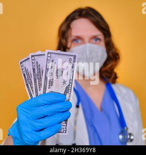 Ein Arzt hält ein Gehalt in US-Dollar. Eine Krankenschwester mit Geld für die Arbeit und einer Schutzmaske auf gelbem Hintergrund Stockfoto