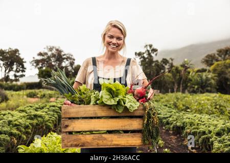 Fröhliche Bio-Farmerin hält eine Schachtel voller frischer Produkte auf ihrem Bauernhof. Glückliche junge Frau lächelt der Kamera zu, während sie in ihrer Gemüsegarde steht Stockfoto
